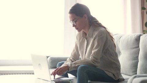 Serious young woman freelancer working on freelance from home typing email on laptop, focused girl using computer for study online at home sitting on couch, female user busy on distance internet job
