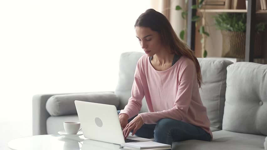 Serious happy young woman typing on laptop browsing chatting in internet use app sit on sofa, smiling focused millennial girl working studying on laptop surfing web looking at computer screen at home Royalty-Free Stock Footage #1028900243