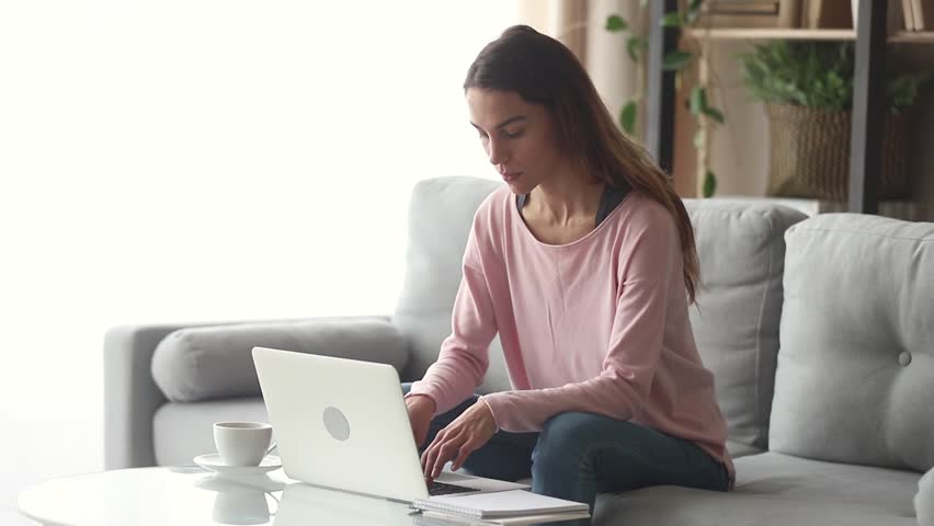 Serious happy young woman typing on laptop browsing chatting in internet use app sit on sofa, smiling focused millennial girl working studying on laptop surfing web looking at computer screen at home | Shutterstock HD Video #1028900243