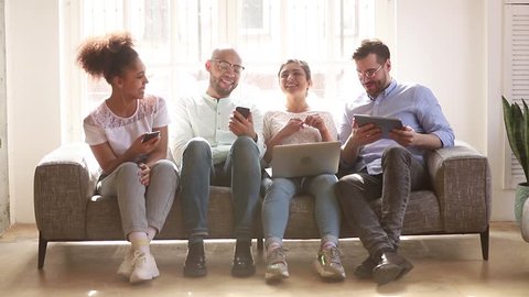 Happy diverse friends group using devices phones laptop digital tablet sit on sofa indoor, cheerful multicultural young people students laughing talking having fun with gadgets technology together