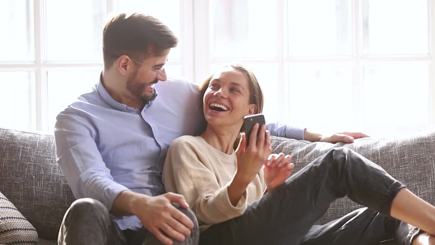 Happy millennial couple talk laugh looking at smartphone using funny apps sit on couch, smiling man and woman relaxing at home having fun in social media on cellphone online watching video on phone Royalty-Free Stock Footage #1028900318