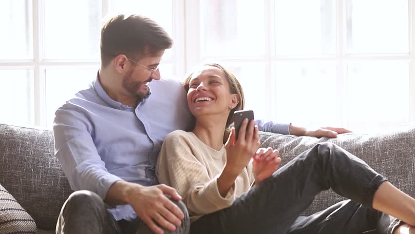 Happy millennial couple talk laugh looking at smartphone using funny apps sit on couch, smiling man and woman relaxing at home having fun in social media on cellphone online watching video on phone | Shutterstock HD Video #1028900318