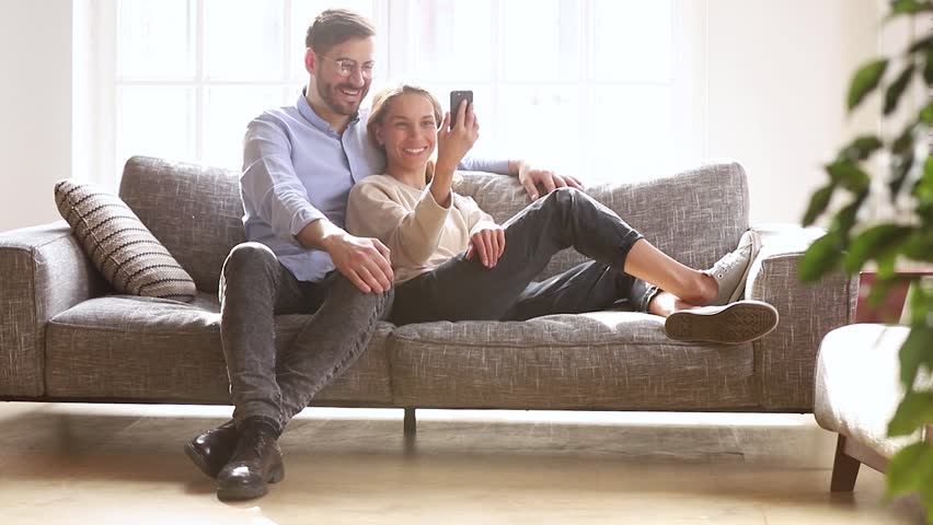 Happy young couple relaxing laughing holding phone talking discuss online news watching videos, millennial girl showing man funny mobile apps having fun with cellphone sit together on sofa at home Royalty-Free Stock Footage #1028900345