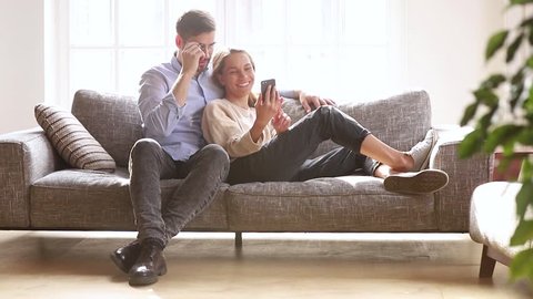 Happy young couple relaxing laughing holding phone talking discuss online news watching videos, millennial girl showing man funny mobile apps having fun with cellphone sit together on sofa at home