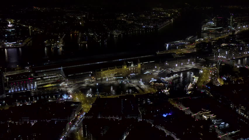 Aerial view of Amsterdam city central night skyline in Netherlands. Flying around Amsterdam Central Station train hub with dark streets and lights, over IJ water in the Dutch province of North Holland