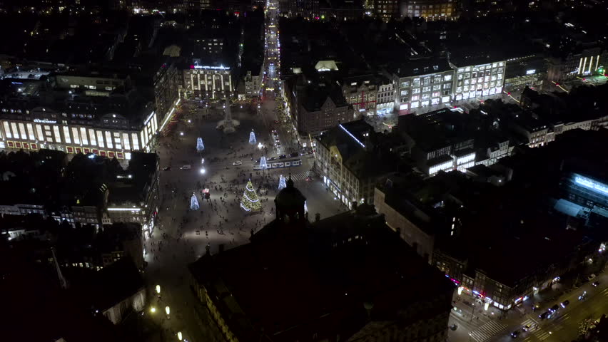 Flying over iconic Dam Square in Amsterdam at night. Famous town square in capital of the Netherlands is home to tourist attractions like National Monument and Royal Palace ft aerial Christmas lights