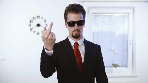 Businessman with glasses shows the fuck you middle finger 4K. Medium shot of a male person in focus dressed up nicely with a red tie. Wall with clock and window in the background.