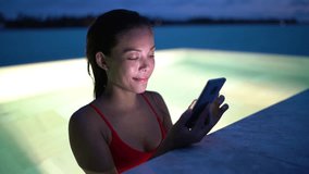 Smartphone - woman using mobile cell phone app on vacation in pool at night on travel holidays. Girl using smartphone app looking at screen smiling happy. Screen light on face.