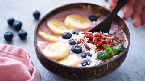 Eating healthy food acai smoothie bowl topped with banana, goji berries, linseeds, blueberries and coconut flakes. Closeup view, slow motion