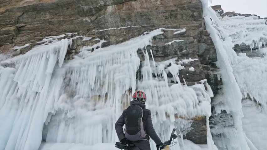 Man is walking beside bicycle near the ice grotto. The rock with ice caves and icicles is very beautiful. The cyclist is dressed in gray down jacket, cycling backpack and helmet. The tires on covered