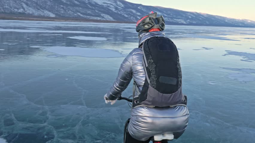 Woman is riding bicycle on the ice. The girl is dressed in a silvery down jacket, cycling backpack and helmet. Ice of the frozen Lake Baikal. The tires on the bicycle are covered with special spikes