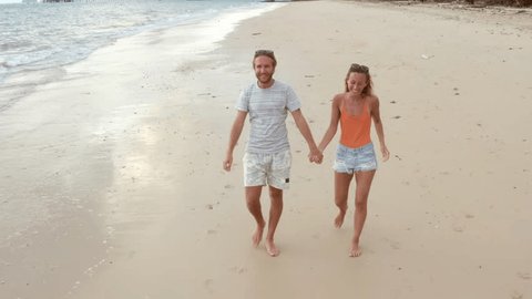 Young couple having a walk on empty tropical beach at sunset. Tracking aerial shot of young couple on vacation having fun on the beach 