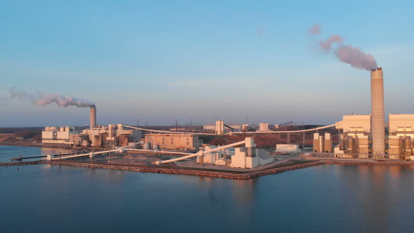 Aerial drone view of a power station on the coast of Lake Michigan at sunrise Milwaukee Wisconsin. Beautiful golden sunrise light illuminates the industrial power plant with its smokestacks cooling towers and structures right on the lakeshore. Crisp 4K footage captures dramatic aerial perspectives of the facility steam emissions the vast expanse of Lake Michigan and the Milwaukee metropolitan coastline. Highlights the contrast between heavy industry and the natural beauty of the Great Lakes at dawn. Perfect for industrial stock footage energy sector videos environmental documentaries sunrise time lapse projects Wisconsin travel or news content about infrastructure and energy.
