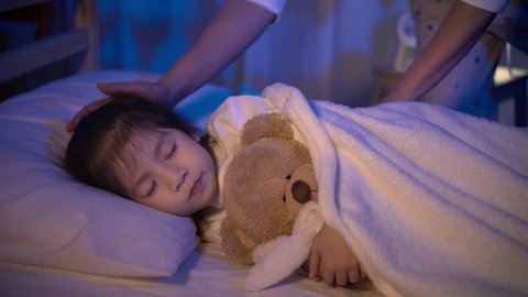 Mother putting blanket on and stroking head for her little girl daughter on bed in a dark bedroom at night, Child asian girl hug teddy bear, Comfortable children at home concept
