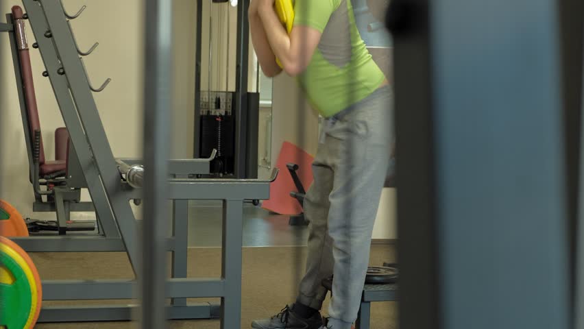 A mature overweight man in a green T-shirt does squats with a weight for a barbell. Fitness training. Healthy lifestyle concept