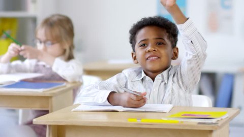 Medium shot of cute little African-American schoolboy sitting at his desk and writing in exercise book, then raising hand and answering question in class – Video có sẵn