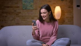 Closeup shoot of young pretty brunette caucasian female having a video call on the phone and smiling sitting on the couch in a cozy apartment indoors