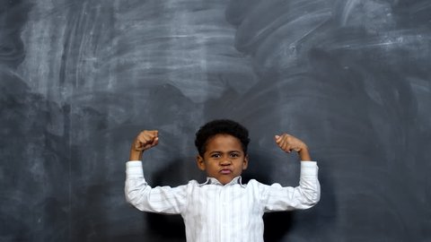 Portrait shot of cute little African-American boy standing against blackboard and showing his biceps