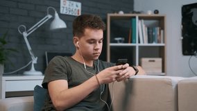 Nice teenage boy is sitting on a couch and listening to podcast in his earphones while texting his friend
