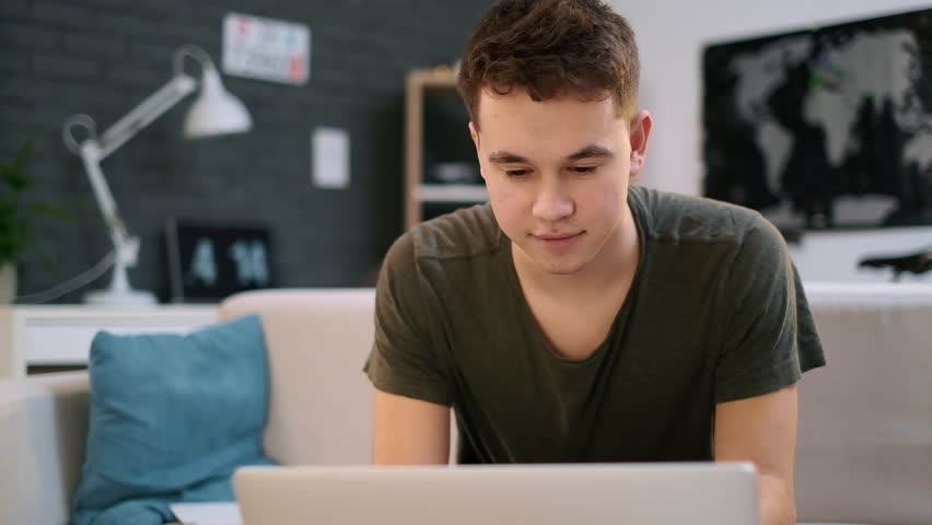 A teenage caucasian boy is happy working on the laptop in his room and nods his head | Shutterstock HD Video #1028962232