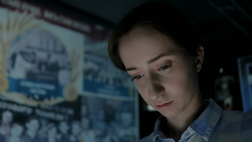Young woman looking at interactive display in multimedia historical museum. Light color from electronic kiosk changing on female face. Evening time, low light. Education and entertainment concept
