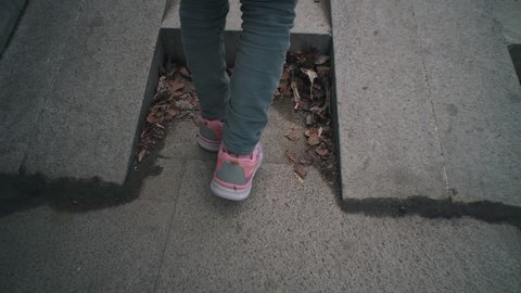Handheld shoot close up of pink sneakers on children's feet climbs stone steps in dry leaves. Child walks on gray stairways in city park.
