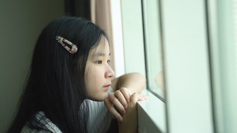 Young asian millennial girl feeling lonely and looking out of window, self quarantine during Covid-19 and coronavirus pandemic