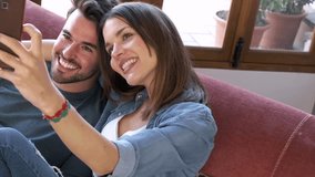 Video of happy young couple taking a selfie with smart phone while sitting on sofa at home.