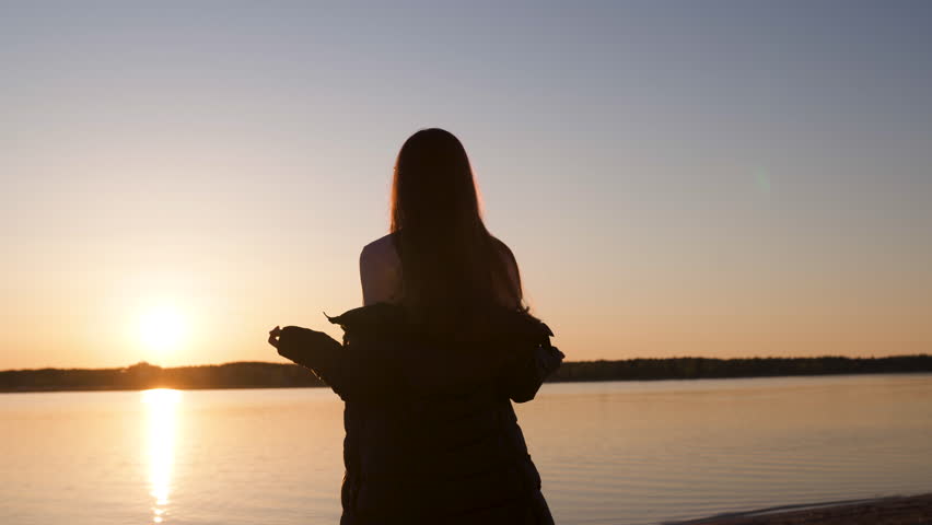 Young woman enjoys sunset during Golden Hour on a river beach in Spring wearing white pants, sweatshort and jacket - Caucasian white girl is a happy traveler
