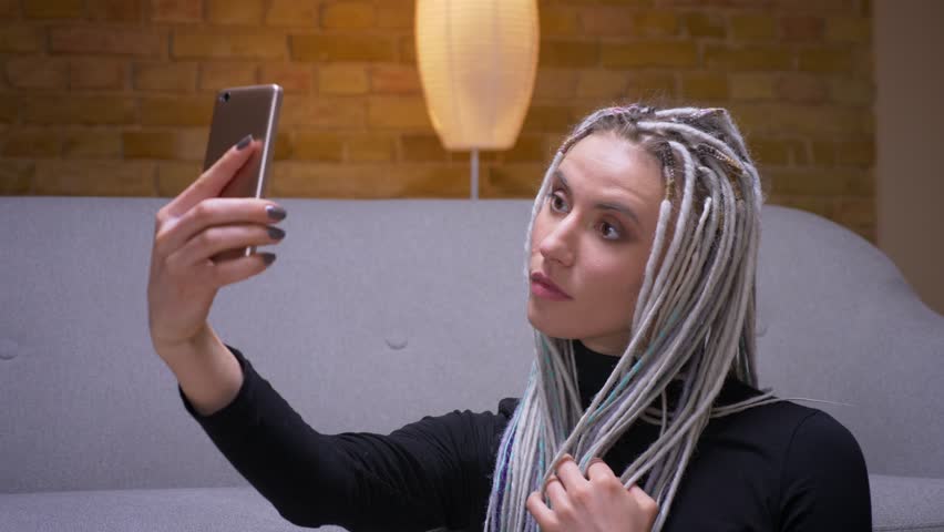 Closeup shoot of young attractive caucasian female with blonde dreadlocks taking selfies on the phone sitting on the floor indoors