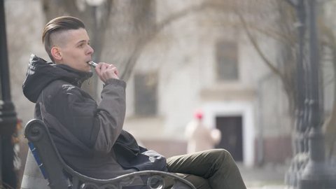  Striking profile of a young man with a short haircut  sitting on a bench, smoking e-cigarette and breathing out dense smoke in spring