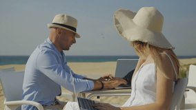 Young couple using laptops on beach. Young man and woman in hats talking and working with laptop computers on sandy beach. Technology concept