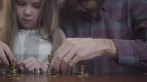 Unrecognizable man in shirt and little concentrated girl sitting at the table counting coins close up. Father and daughter making stacks from coins. The dad teaching kid economy. Money saving concept