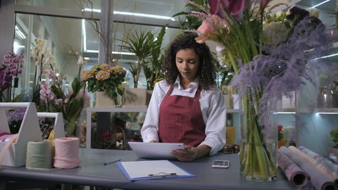 Mixed race female florist looking at tablet pc display accepting orders from customers on delivery of flowers. Modern floristical salon provides flower delivery service, employee networking at counter