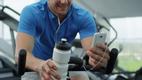 Close up shot of middle-aged Caucasian man using exercise bike in gym, drinking water from bottle and watching videos on smartphone