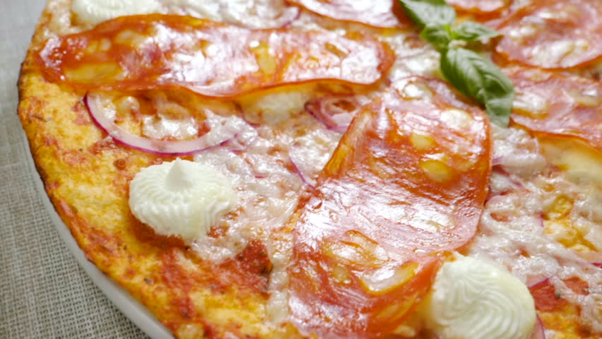 Italian food. Close-up shot of delicious pizza with tomato sauce, salami slicing, fresh basil leaves, parmesan and cream cheese, red onion served on a round white plate. Slow motion. HD