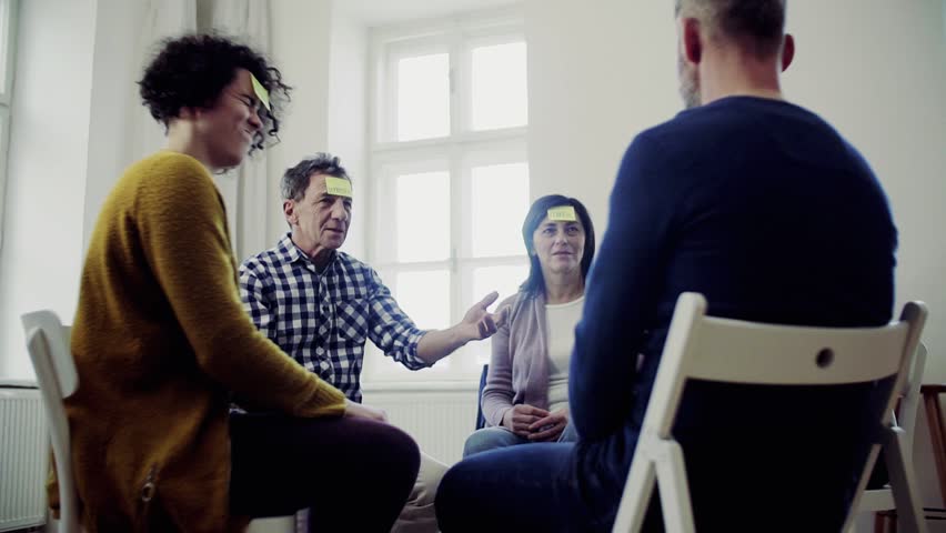 Men and women sitting in a circle during group therapy, adhesive notes on forehead. | Shutterstock HD Video #1029014843