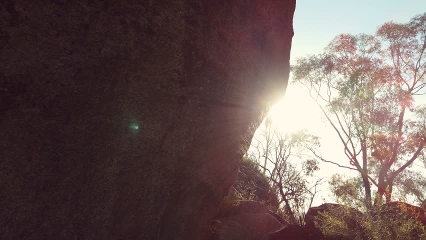 Glide shot of bright sunlight shining past a large rock with gum trees in the background in the Australian bush land.
