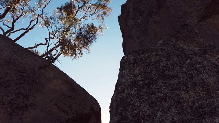 Glide shot of bright sunrays shining past a large rock with gum trees in the background in the Australian bushland.