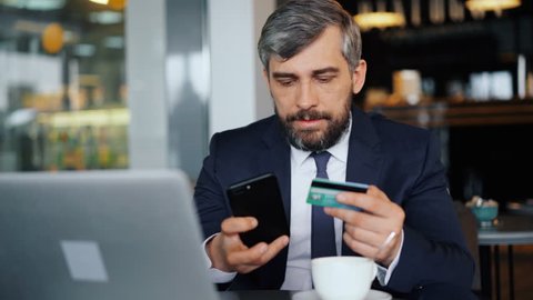 Businessman is making online payment holding credit card and smartphone sitting in cafe then smiling after successful transaction. Modern technology and banking concept.