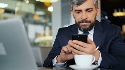 Bearded office worker in suit is using smartphone during coffee break touching screen and smiling sitting at table in cafe. Technology and lunch time concept.