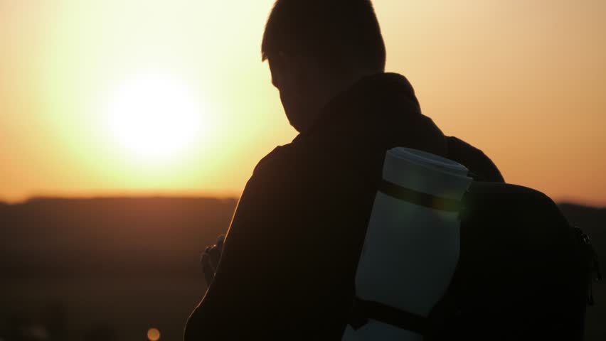 A silhouette young tourist man with a backpack uses a mobile phone. In the rays of the setting sun. Slow motion. Travel concept. Always online.
