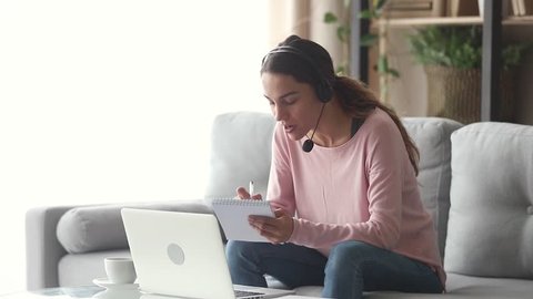 Smiling young woman interpreter wearing headset talking on making notes, happy female online teacher tutoring teaching by distant video call, student e learning on web sitting on sofa at home