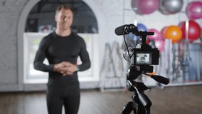 Male fitness coach waving, speaking and showing muscles to camera while recording video in gym for sports vlog; focus in the foreground