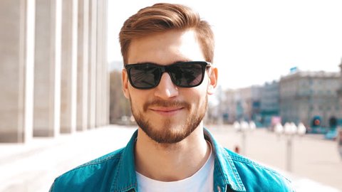 Portrait of handsome smiling stylish hipster lumbersexual model. Man dressed in jeans jacket clothes. Fashion male posing on the street background in sunglasses. Slow motion