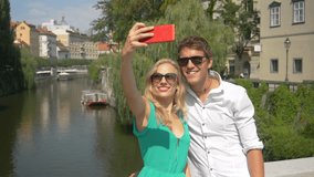 SLOW MOTION, CLOSE UP: Cheerful young travelers take selfies from a bridge in sunny Ljubljana. Handsome Caucasian man gives the thumbs up while filming a video blog with his cute blonde girlfriend.