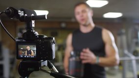 Rack focused shot of professional fitness coach looking at camera and telling about bodybuilding supplements while recording online commercial in gym