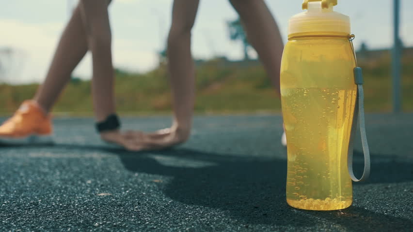 Close up of sportsman water bottle on the outdoor stadium floor in the front and stretching femae in the back