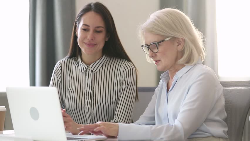 Older female professional mentor teach young intern worker learn new skill look at laptop, mature businesswoman executive manager explain colleague computer work talk consult client with computer Royalty-Free Stock Footage #1029051434