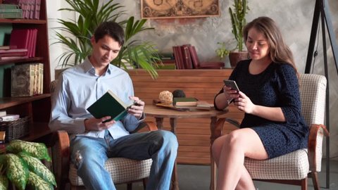 The guy and the girl are sitting in the home library, a man in a blue shirt and jeans, a girl in a dark dress. The girl looks into the phone, the guy reads a book.
